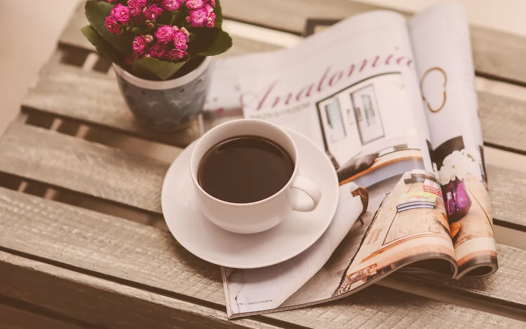 A photo of a white coffee cup sitting on a Woden table with a fold magazine and pink flower pot